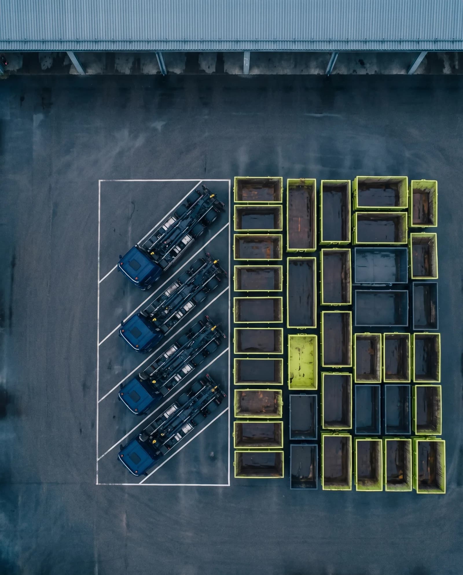 Four navy lorries parked in a row beside a grid of lime and grey containers, seen from directly above